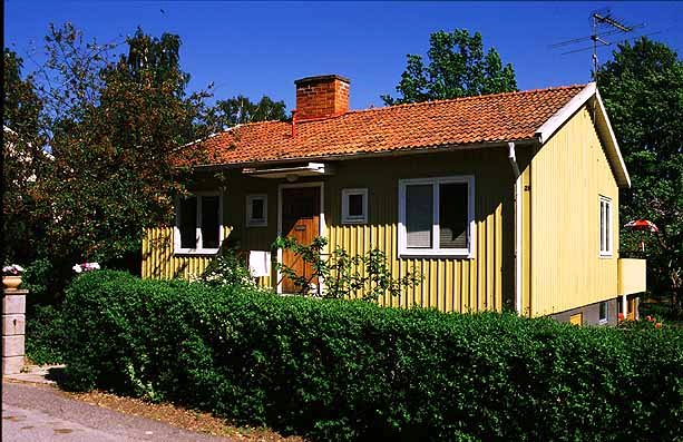 Tidstypisk exteriör med gul locklistpanel, rött tegeltak, tvåluftsfönster och litet skärmtak. Skogst